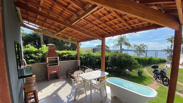 An outdoor patio with a barbecue grill, a table with chairs, a white bathtub, and a view of a lake and palm trees. - Villa Querência (Avare)