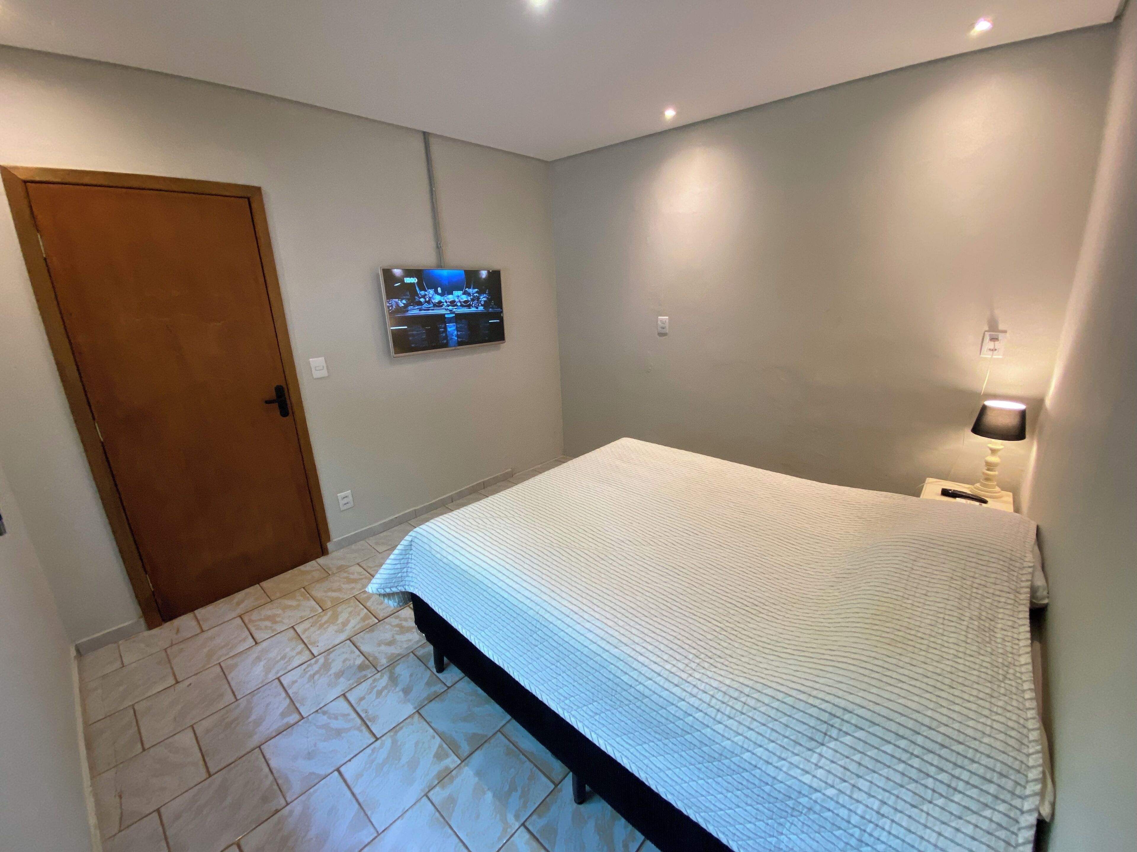 A bedroom with a bed, a TV mounted on the wall, a door, and a nightstand with a lamp.