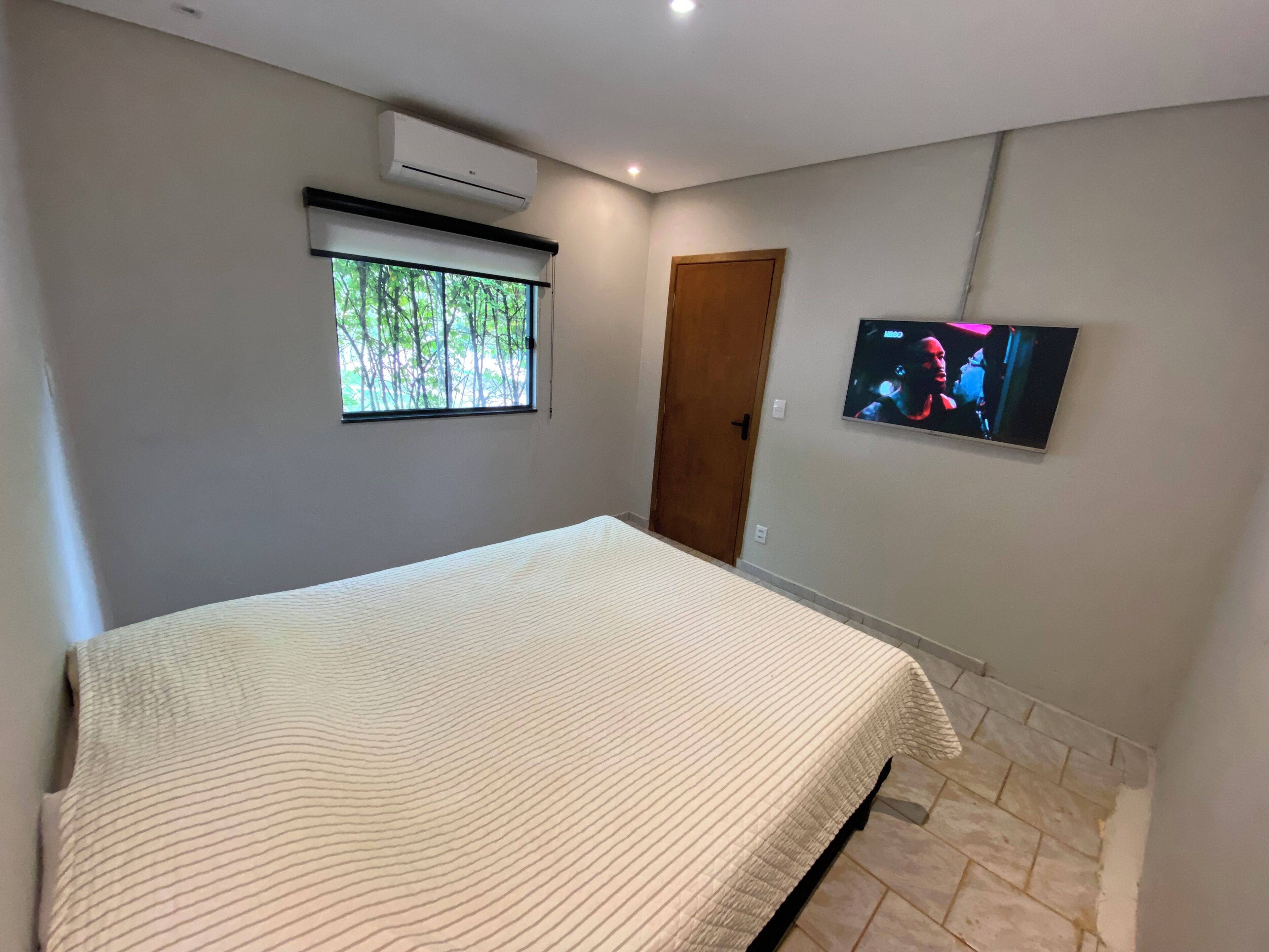 Un dormitorio con cama, aire acondicionado, ventana con vista a árboles, una puerta y televisor en la pared.