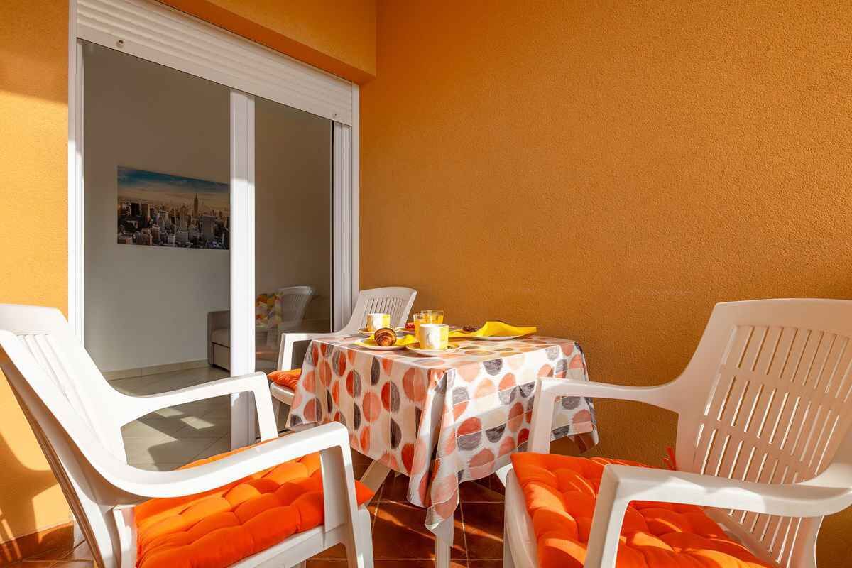 A balcony with a table set for two, white plastic chairs with orange cushions, and a cityscape view through a glass door.