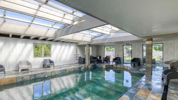An indoor swimming pool with clear water, surrounded by stone tiles, and large windows allowing natural light. - Le Paradis (Orcival)