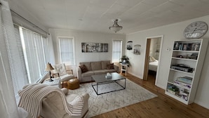 A living room with a sofa, coffee table, and bookshelf, featuring a ceiling fan, window blinds, and a clock. - Waterfront 3 Bedroom cottage located in beautiful Turkey Point (Vittoria)