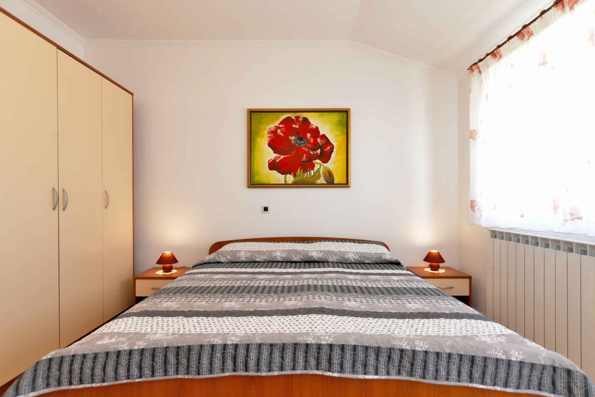 Ein Schlafzimmer mit Bett, Nachttisch, Lampe, Kleiderschrank und einem Gemälde einer roten Blume an der Wand.