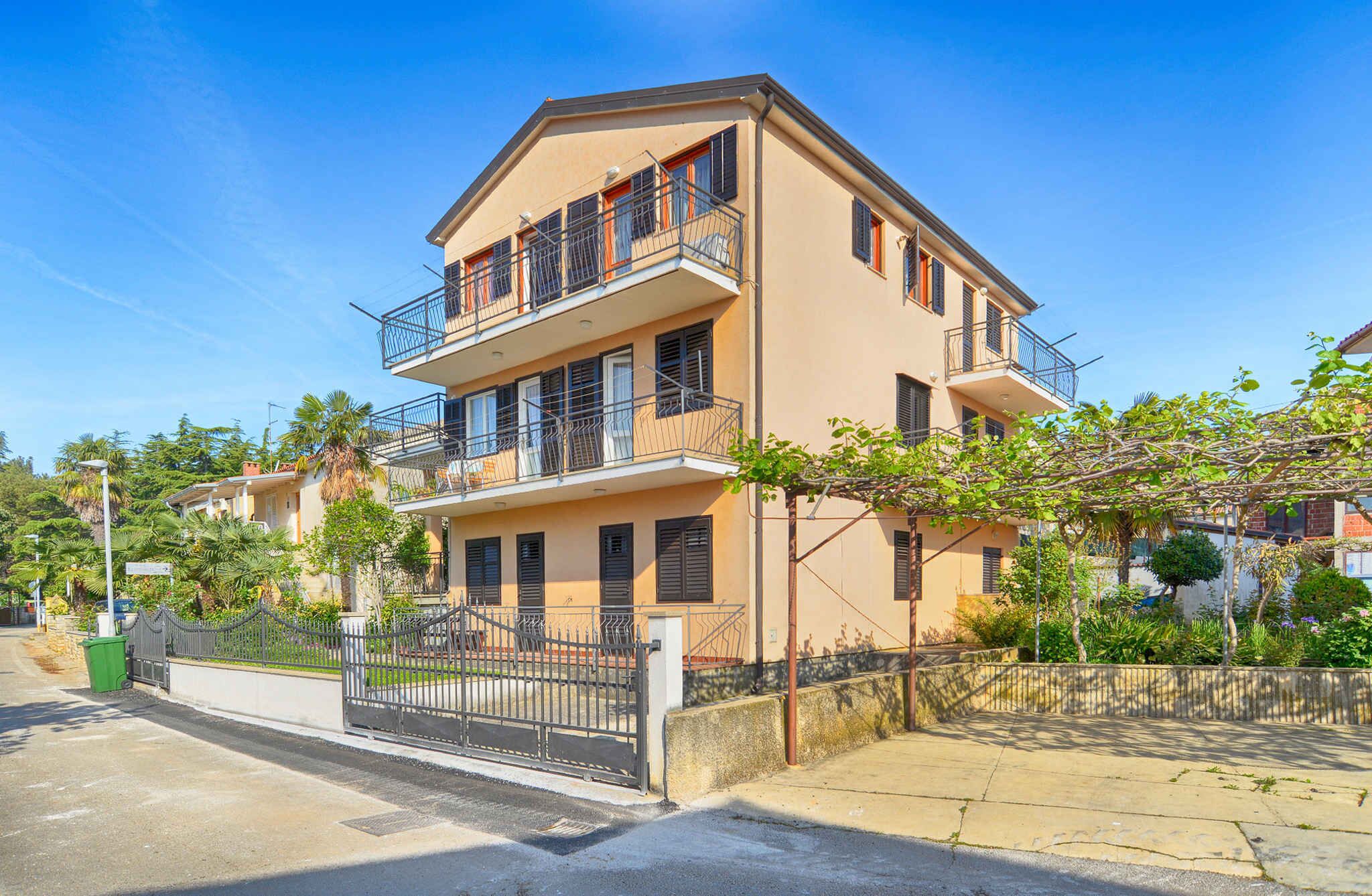 A two-story residential building with a balcony on the upper floor, a gated entrance, and surrounding greenery.