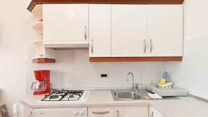 Cocina compacta con gabinetes blancos, estufa a gas, cafetera y pileta.