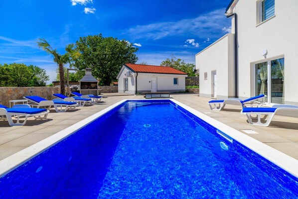Une piscine rectangulaire à l’eau bleue, entourée de chaises longues blanches avec des coussins bleus. On aperçoit, au fond, un petit bâtiment avec un toit rouge et une cheminée.