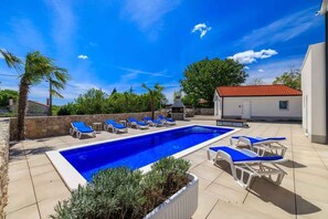 Une piscine rectangulaire à l’eau bleue, entourée de chaises longues et d’un bâtiment blanc doté d’un toit rouge.