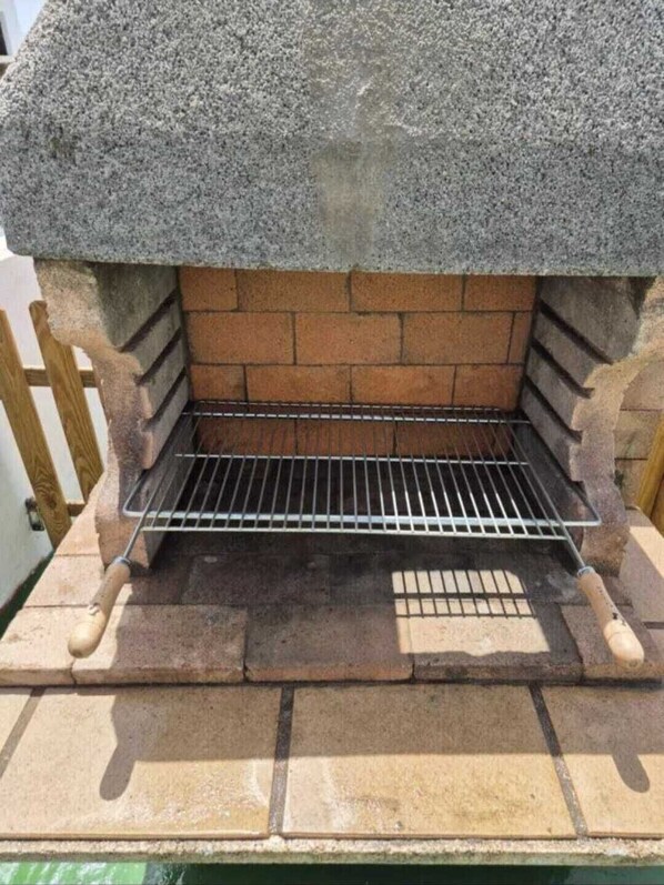 A stone barbecue grill with a metal rack, set on a brick base with a tiled floor.