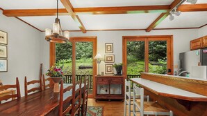 A wooden dining table with chairs, a kitchen area with a refrigerator, and a view of greenery through large windows. - Lush, Private, & Chic Blue Ridge Retreat with 2 Fireplaces- 3 BR - 2.5 BA (Boone)