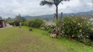 A residential area with a brick house, a covered patio, and a lush garden with pink flowers and a palm tree. - Suite Gold Rocks Glamping (El Penol)