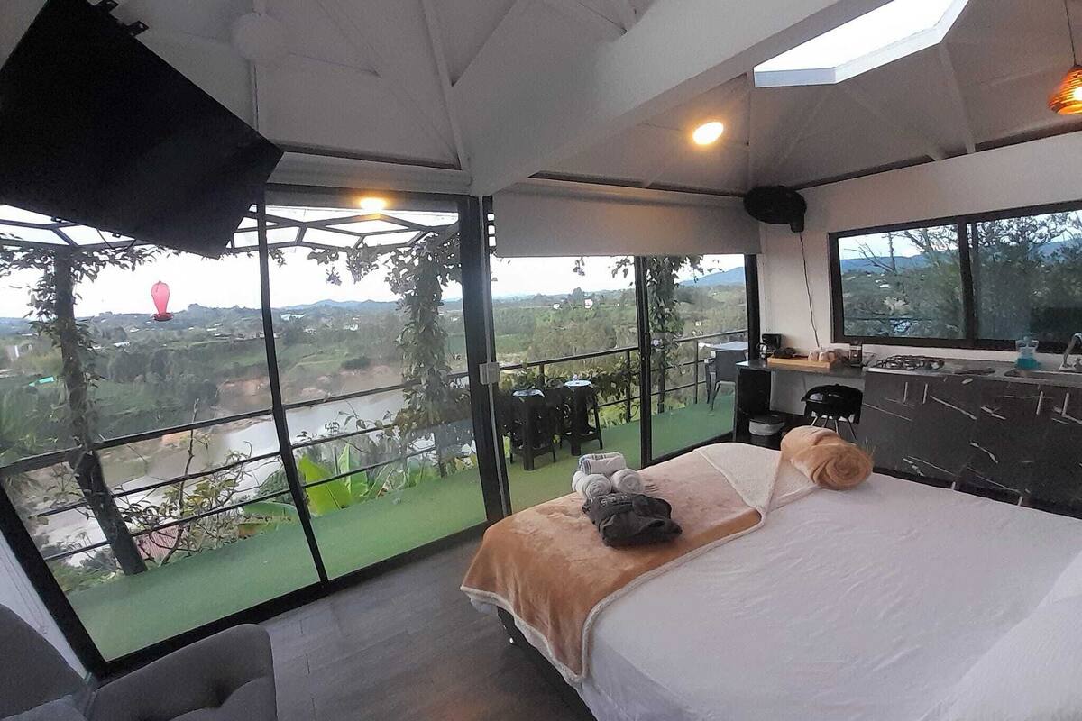 A modern bedroom with a large bed, a small seating area, and a balcony with a view of the countryside.