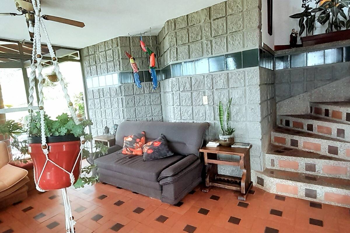 A living room with a sofa, a coffee table, and a hanging plant. There is a brick fireplace and a staircase with a railing.