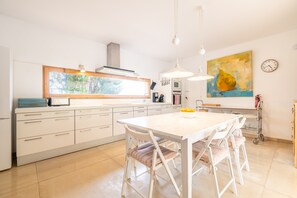 A modern kitchen with white cabinetry, a large window, pendant lights, and a dining area with white chairs.