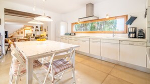 Moderni keittiö, jossa on valkoiset kaapit, suuri ikkuna sekä ruokailutila, jossa on valkoinen pöytä ja tuolit.