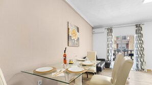Un coin repas avec une table en verre, des chaises blanches, un tableau au mur et une vue sur les bâtiments par la fenêtre.