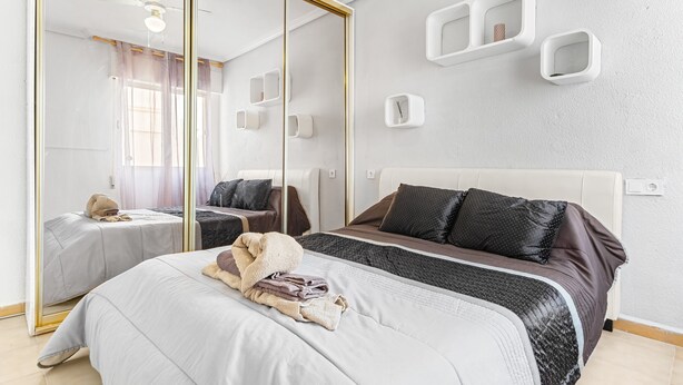 Une chambre d’hôtel moderne avec un grand miroir, un lit recouvert d’une courtepointe blanche et marron, une table de chevet avec une lampe et une étagère fixée au mur.