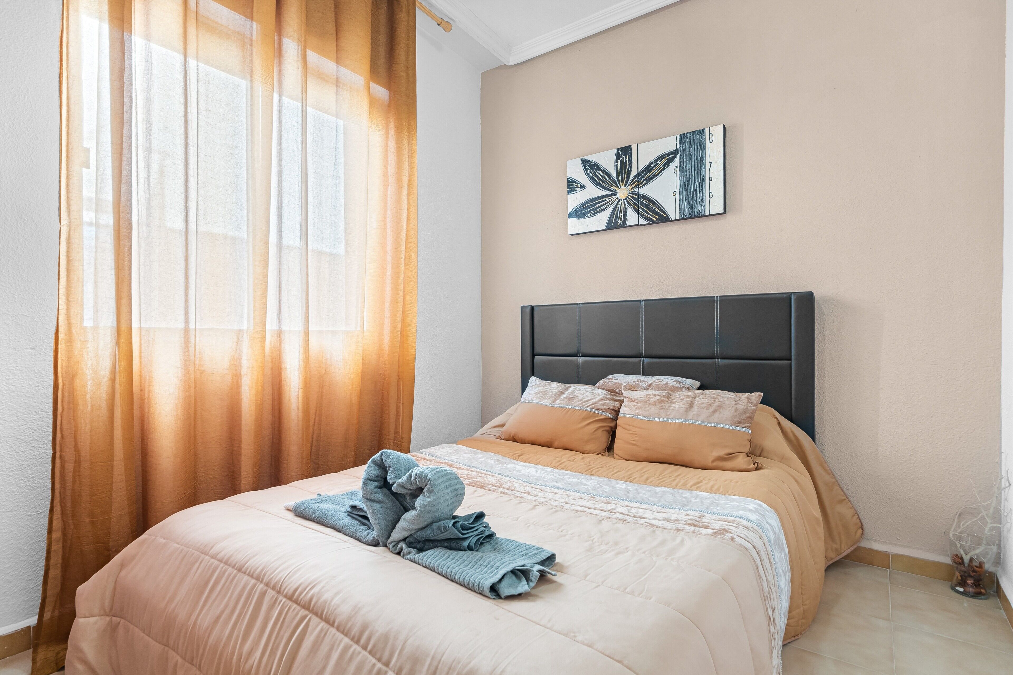 Una cama bien hecha con cabecera, una ventana con cortinas translúcidas y una obra de arte en la pared.