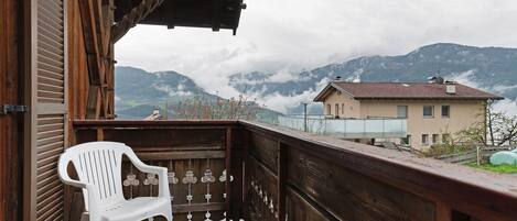 A wooden balcony with a white plastic chair, overlooking a mountain landscape with clouds.