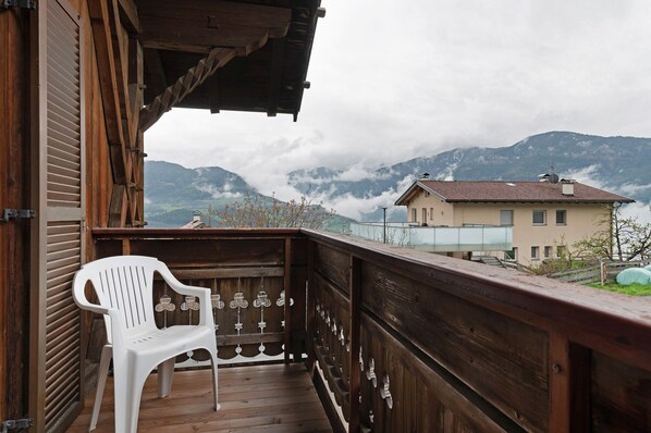 A wooden balcony with a white plastic chair, overlooking a mountain landscape with clouds.
