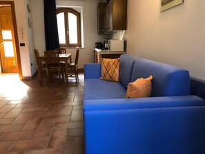 Un sofá azul con un cojín estampado, una mesa de comedor de madera con sillas, y una zona de cocina con armarios y un microondas.