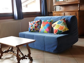 A blue sofa with colorful geometric pillows, a wooden table, and a wooden cabinet in a room with tiled flooring and a window with curtains.