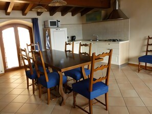 A dining area with a wooden table and blue upholstered chairs, a kitchen with white cabinets, and a large window with curtains.