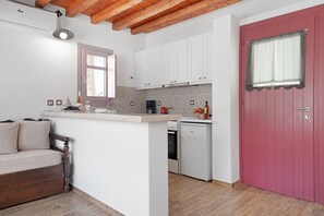 Een kleine keuken met witte kasten, een rode deur, een houten plafond en een eethoekje met een bankje.