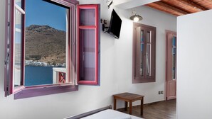 A bedroom with a bed, a small wooden table, a TV mounted on the wall, and a view of a mountainous landscape through an open window. - Apartment 'Nicolas House 1' with Sea View, Wi-Fi and Air Conditioning (Rakhídhion)