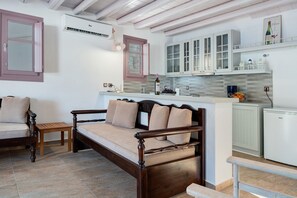 A bedroom with a wooden bed, a small bedside table, a chair, and a kitchenette with cabinets and a refrigerator.