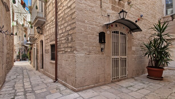 A narrow cobblestone street with stone buildings, a potted plant, and a metal gate. - Apartment 'Home Sweet Home' with Balcony, Wi-Fi and Air Conditioning (Giovinazzo)