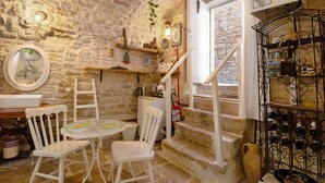 Un interior rústico de piedra con una mesa redonda, sillas blancas, una estantería para vinos y una escalera.