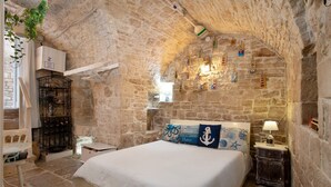 Un dormitorio de paredes de piedra con una cama, una mesita, una silla y una estantería con adornos.
