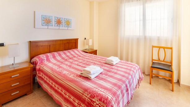 A room with a bed, a wooden chair, a nightstand, a lamp, and a window with curtains.