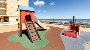 A playground with a slide and climbing structure, a beach view, and a building in the background.