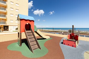 Um parquinho com escorrega e estrutura de escalada, vista para a praia e um edifício ao fundo.