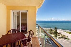 A balcony with a table and chairs, a sliding glass door, and a view of the sea and a road.