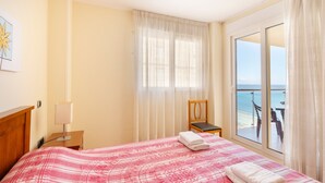 A hotel room with a bed, a chair, a nightstand, a lamp, and a view of the sea through a large window.