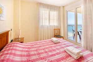 Quarto de hotel com cama, poltrona, mesa de cabeceira, luminária e vista para o mar através de uma grande janela.