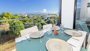 Una mesa de comedor dispuesta con platos, vasos y cubiertos, con vistas a un paisaje pintoresco con árboles y edificios.
