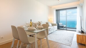 Ein moderner Essbereich mit einem Glastisch, weißen Stühlen und einer Weinflasche. Im Hintergrund sind eine Couch und ein Sessel zu sehen, mit Blick auf das Meer durch große Fenster.