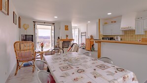 Uma área de jantar com mesa posta para quatro, uma cadeira de vime e uma cozinha com armários brancos e painel de azulejos.