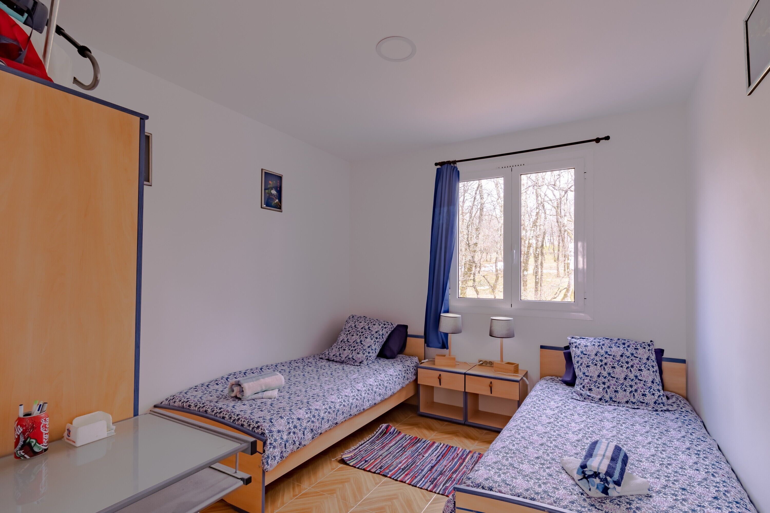 A room with two single beds, a desk, a chair, a wardrobe, and a window with curtains.
