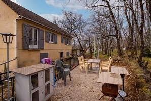 Un bâtiment à la façade jaune, doté d’un balcon avec des volets gris, d’un espace extérieur avec des chaises en plastique et d’un sol recouvert de gravier.