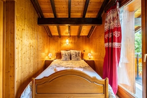 A wooden room with a bed, two bedside lamps, and a window with a red curtain.