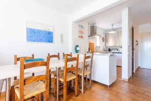 Ein Essbereich mit einem weißen Tisch und Holzstühlen, im Hintergrund eine Küche und ein gefliester Boden.