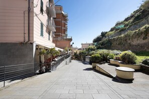 A paved pathway leading to a building with a balcony, surrounded by greenery and a hillside.
