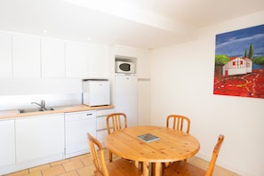 A compact kitchen with white cabinets, a microwave, and a dishwasher. There's a wooden dining table with chairs. A painting of a house is on the wall.