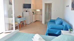 A compact room with a bed, a small table with a bowl of fruit, a kitchenette, and a shower area.