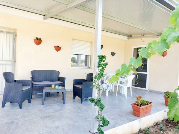 A covered outdoor seating area with wicker furniture, potted plants, and a view of the interior.