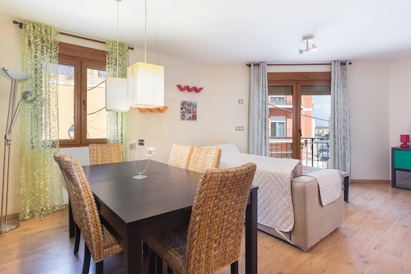 A room with a dining table, chairs, a bed, a lamp, and a window with curtains.